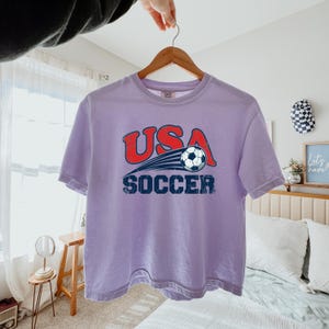 May include: A light purple short-sleeve t-shirt featuring a graphic print with the red text USA above a black and white soccer ball with motion lines, and the blue text SOCCER below. The design has a distressed, weathered appearance.