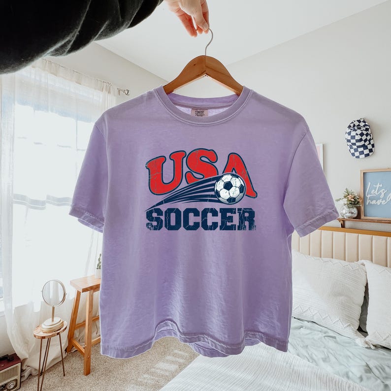 May include: A light purple short-sleeve t-shirt featuring a graphic print with the red text USA above a black and white soccer ball with motion lines, and the blue text SOCCER below. The design has a distressed, weathered appearance.