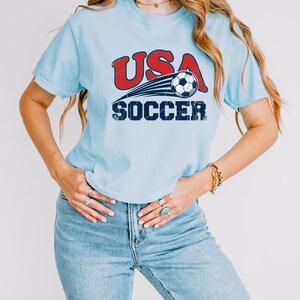 May include: A light blue short-sleeve t-shirt featuring a graphic print with the red text USA above a black and white soccer ball with motion lines, and the blue text SOCCER below. The design has a distressed, weathered texture throughout.