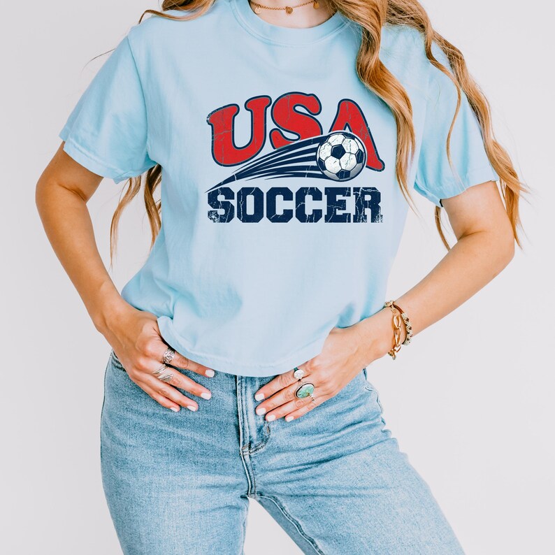 May include: A light blue short-sleeve t-shirt featuring a graphic print with the red text USA above a black and white soccer ball with motion lines, and the blue text SOCCER below. The design has a distressed, weathered texture throughout.