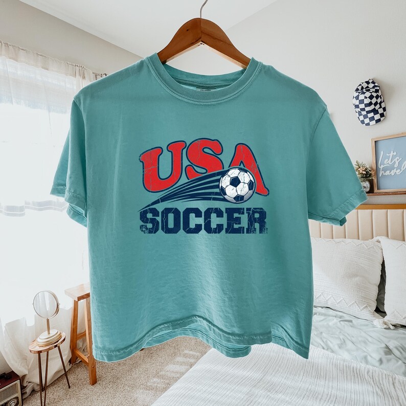 May include: A teal short-sleeve t-shirt featuring a distressed graphic print on the chest. The design includes the red text USA, a black and white soccer ball with motion lines, and the dark blue text SOCCER positioned below the ball.