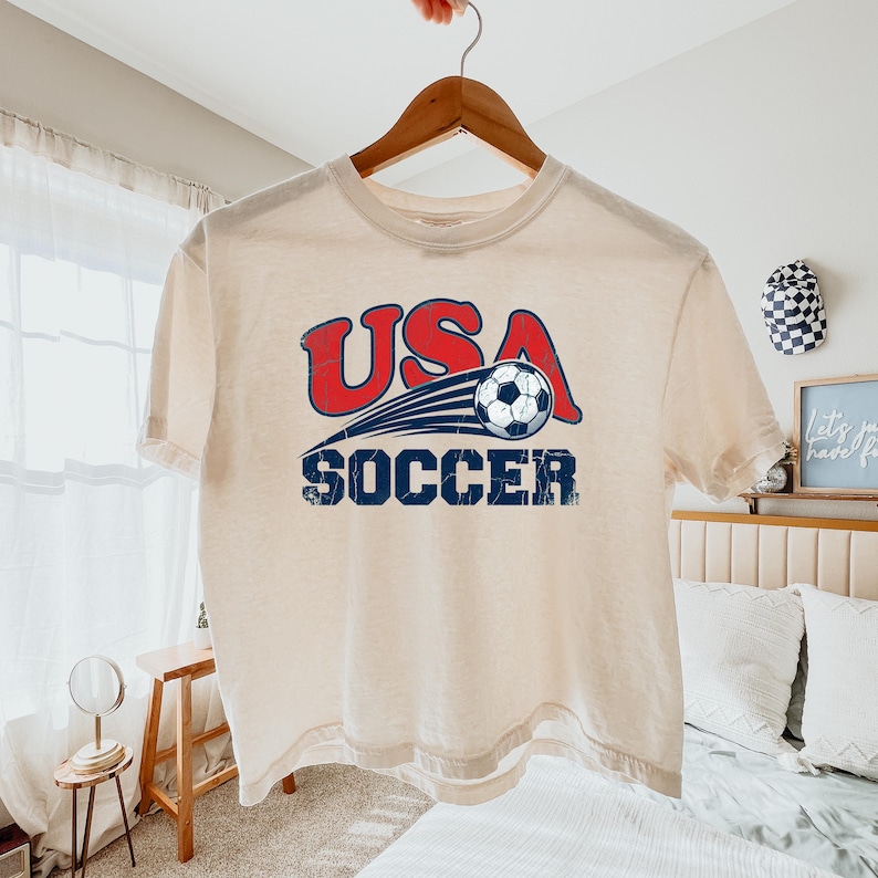 May include: A cream-colored short-sleeve t-shirt featuring a graphic print with the red text USA above a blue soccer ball in motion, followed by the blue text SOCCER. The design has a distressed, weathered texture throughout the lettering.