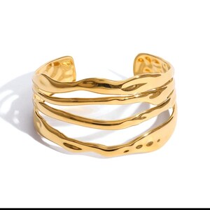 Peut inclure: Un bracelet manchette doré avec un design texturé et ondulé. Le bracelet ouvert présente trois bandes parallèles et fluides. Le bijou est posé sur un fond blanc, soulignant son éclat métallique.