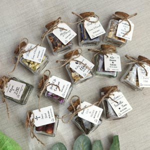 May include: A collection of small square glass jars filled with various dried tea leaves and botanicals. Each jar features a cork stopper, a brown twine tie, and a white paper tag printed with names, dates, and tea types for personalized gifts.