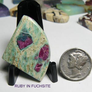 May include: A polished, triangular-shaped ruby in fuchsite gemstone cabochon featuring a mottled mint green and cream matrix with embedded deep magenta-red ruby inclusions, displayed on a black stand next to a silver 1941 Mercury dime for scale.