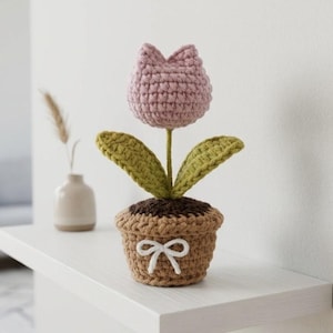 May include: A crocheted flower in a pot featuring a dusty pink tulip-shaped bloom, two textured olive green leaves, and a brown pot with a dark brown top rim. A small white yarn bow is tied around the centre of the light brown crocheted pot.