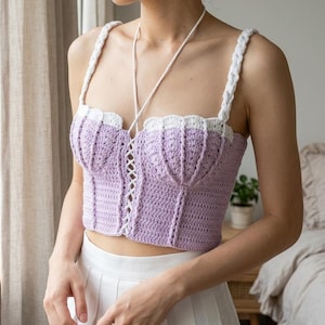 Pode incluir: Um top de crochê lavanda com acabamentos e alças brancas. O top apresenta uma frente estilo espartilho com fecho de cadarço. As alças são trançadas. O top é combinado com uma saia plissada branca.