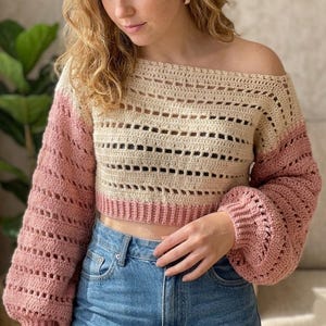May include: A cream and pink crochet off-the-shoulder sweater. The sweater has long, puffy sleeves in a solid pink colour. The body of the sweater is cream-coloured with a rectangular openwork design. The bottom hem and cuffs are a solid pink.
