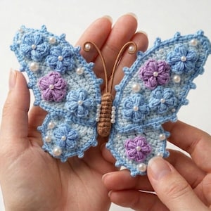 May include: A handmade crochet butterfly in shades of light blue, lavender, and brown. The butterfly wings are adorned with small blue and lavender flowers, and white pearl beads. The butterfly is held in a person's hands.