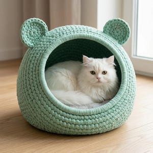 May include: A mint green, crocheted cat bed shaped like a bear's head. A fluffy white cat rests inside the bed. The bed has a round shape with two small, rounded ears on top. The cat bed is on a wooden floor.