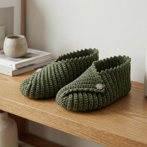 Peut inclure: Une paire de chaussons en crochet vert olive avec une texture côtelée, un bord supérieur festonné et un bouton décoratif en cristal argenté sur le côté extérieur de chaque chausson, offrant un ajustement structuré et sécurisé.