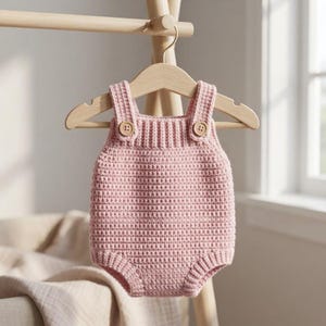 May include: A light pink crocheted romper featuring a textured stitch pattern, a ribbed neckline, and two small wooden buttons on the front straps. The garment is displayed on a light-coloured wooden hanger, showcasing its simple and functional design.