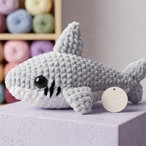 May include: A gray and white crocheted shark plush toy with black eyes and black markings. The shark has a tag attached. The background includes a shelf with colorful yarn balls.