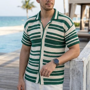 Può includere: Una camicia all'uncinetto a maniche corte con righe orizzontali color crema e verde. La camicia ha un colletto e una chiusura con bottoni. La persona indossa un orologio nero e pantaloni bianchi. Scena di spiaggia sullo sfondo.