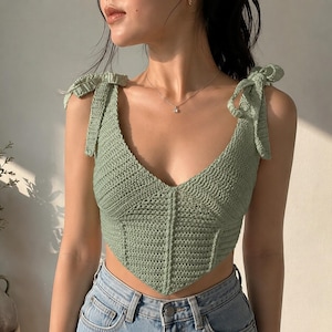 May include: A sage green crochet crop top featuring a V-neckline, a pointed hem, and shoulder straps that tie into bows. The garment is constructed with a textured stitch pattern and is paired with light blue denim jeans with a visible metal button.