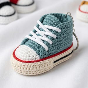 Könnte beinhalten: Ein gehäkelter Sneaker-Schlüsselanhänger in Blaugrün mit weißen Schnürsenkeln, weißer Zehenkappe und einer cremefarbenen Sohle mit einem dünnen roten Streifen und einer schwarzen Akzentlinie. Der Minischuh zeigt sichtbare Maschen und einen Metallring.