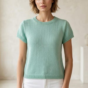 以下が含まれることがあります： 淡い緑色の半袖ニットトップス。クルーネックとリブ編みの裾と袖口が特徴です。生地は繊細な質感です。トップスは無地で、柔らかく通気性の良い素材で作られているようです。