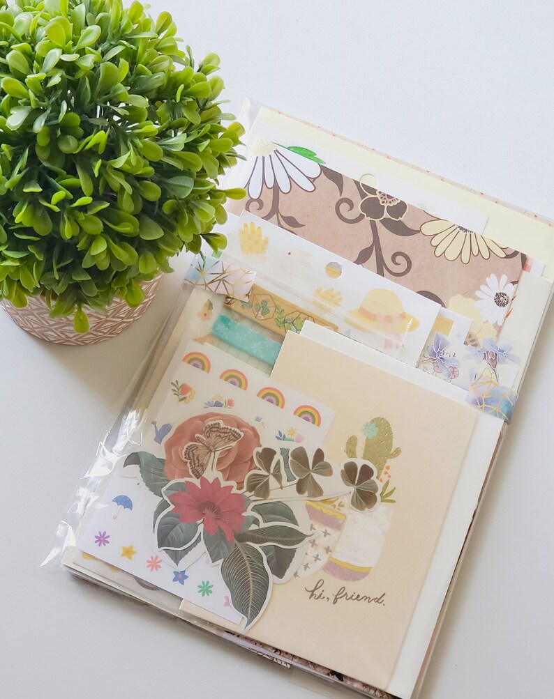 May include: A set of colorful paper ephemera, including patterned paper, stickers, and washi tape. The set features a floral theme with butterflies, clovers, and a cactus. The text "hi, friend." is visible on one of the papers.