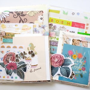 May include: A clear plastic bag filled with colorful paper ephemera, including stickers, washi tape, and patterned paper. The bag is divided into two sections, with one side featuring a floral design and the other side featuring a cactus design. The text "BLOSSOM FRESH" and "Creativity takes courage" is visible on the paper.