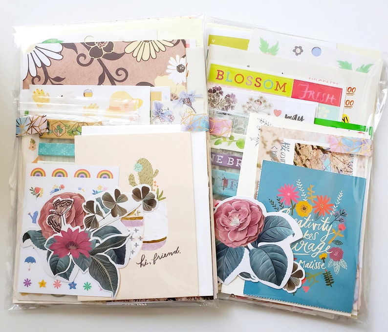 May include: A clear plastic bag filled with colorful paper ephemera, including stickers, washi tape, and patterned paper. The bag is divided into two sections, with one side featuring a floral design and the other side featuring a cactus design. The text "BLOSSOM FRESH" and "Creativity takes courage" is visible on the paper.