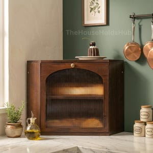 May include: A dark wood cabinet with a ribbed glass door and a small, round brass knob. The cabinet is on a marble countertop, with a small potted plant and a glass bottle of oil in front of it. Copper pans hang on the wall.