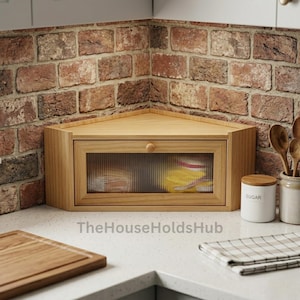 May include: A light-toned wooden corner bread box featuring a rectangular front panel with a fluted glass window and a small round wooden knob. The bread box is designed to fit into a corner and has a smooth, natural wood grain finish throughout.