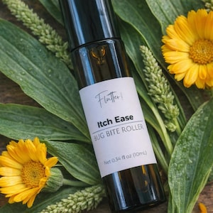 Puede incluir: Un frasco rodillo de vidrio oscuro etiquetado "Flutter: Itch Ease BUG BITE ROLLER" con un peso neto de 10 ml. La botella está rodeada de hojas verdes, flores amarillas y pétalos de flores.