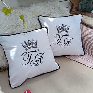 Pode incluir: Duas almofadas decorativas brancas com borda preta. Cada almofada apresenta um desenho de coroa preta acima das letras entrelaçadas "TA". As almofadas estão colocadas numa superfície clara, adequadas para decoração.