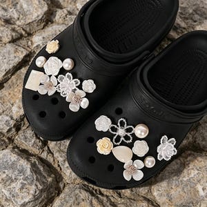 Könnte beinhalten: Schwarze Clogs, verziert mit dekorativen Blumenanhängern und Kunstperlen. Die Anhänger umfassen weiße und cremefarbene Blumen, die dem lässigen Schuhwerk einen Hauch von Eleganz verleihen. Die Clogs werden auf einer strukturierten, felsigen Oberfläche präsentiert.
