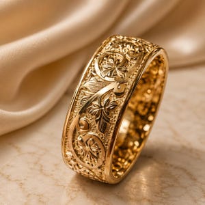 Puede incluir: Un anillo ancho de oro con un intrincado patrón en relieve de enredaderas entrelazadas y motivos florales. La superficie metálica tiene un acabado pulido con detalles texturizados dentro del diseño tallado, creando una apariencia dimensional.