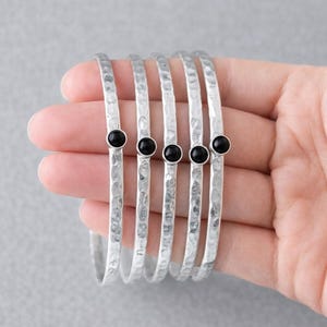 May include: A set of six silver bangles with a hammered texture, each adorned with a small, round black stone. The bracelets are displayed against a soft gray background, showcasing their simple elegance.