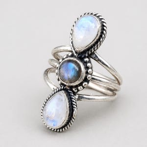 Puede incluir: Un anillo de plata con tres piedras preciosas. Dos piedras lunares en forma de pera flanquean una piedra central redonda e iridiscente. El anillo tiene un diseño multibanda con un borde de cuerda trenzada.