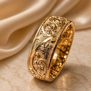 Può includere: Un anello dorato con un intricato motivo floreale. L'anello presenta incisioni dettagliate di fiori, foglie e motivi a spirale. La fascia spessa ha una finitura lucida, che riflette la luce. Lo sfondo è di un colore morbido e neutro.