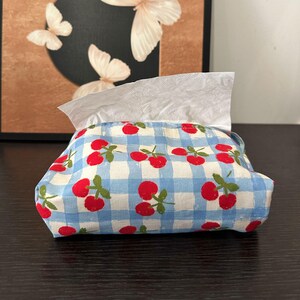 以下が含まれることがあります： 水色と白のギンガムチェック柄の布製ティッシュケース。赤いサクランボと緑の茎が繰り返しプリントされています。長方形のポーチの上部にある開口部から、白いティッシュが1枚引き出されています。