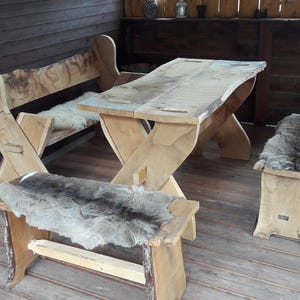 Könnte beinhalten: Ein rustikales Ess-Set für den Außenbereich aus Holz. Das Set umfasst einen Tisch mit X-förmigem Gestell, zwei Bänke und einen Hocker. Jeder Sitz ist mit einem Fell bedeckt. Die Möbel sind aus hellem Holz gefertigt.