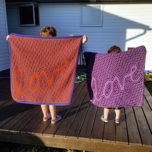 Könnte beinhalten: Zwei Kinder halten gehäkelte Decken hoch, auf denen das Wort "LOVE" in großen Buchstaben geschrieben steht. Die Decke links ist orange und lila, die Decke rechts ist lila und weiß.