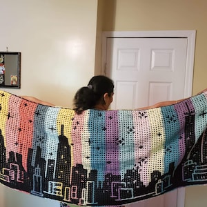 Può includere: Una persona tiene in mano una coperta all'uncinetto colorata con un disegno di città nero. La coperta presenta strisce rosa, gialle, blu, verdi e viola.