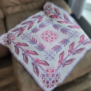 May include: A white crocheted throw blanket with a floral design in shades of pink and purple.