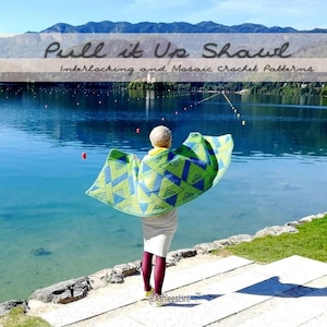 Op de afbeelding: Een vrouw draagt een groen, blauw en geel gehaakt sjaal met een geometrisch patroon. De sjaal heet "Pull it Up Shawl" en heeft in elkaar grijpende en mozaïek haakpatronen. De vrouw staat op een stenen pad aan een meer met bergen op de achtergrond.
