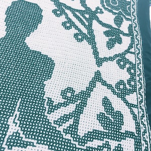 May include: A white and dark green crocheted blanket featuring a repeating geometric border and a central motif of a silhouette figure standing beside a stylized tree with leaves and branches, created using a grid-based stitch pattern.
