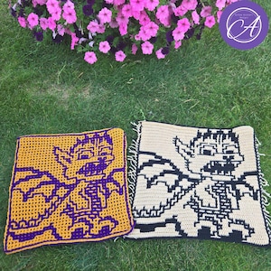 May include: Two crocheted square pillows with pixelated monster designs. One is gold with a purple monster, the other is beige with a black monster. The pillows are on green grass, with pink flowers in the background.