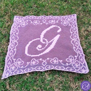 May include: A handmade, crocheted blanket in shades of purple, featuring a large letter "G" in a lighter purple. The blanket has a decorative border with heart and geometric patterns. The blanket is displayed on a bed of green grass.