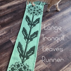 May include: A crocheted green and dark green runner with a floral design. The runner is called "Large Tranquil Leaves Runner" and is by Ashlee Brotzel Designs.