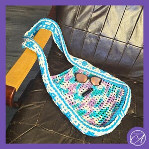 May include: A blue and white crocheted tote bag with a pink and green pattern. The bag is sitting on a brown leather chair with sunglasses and a lipstick inside.