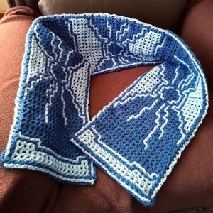 May include: A blue and white crocheted scarf with a geometric pattern. The scarf is folded in half and laid on a brown surface.
