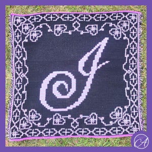 Puede incluir: Un cuadrado de crochet negro y blanco con un borde rosa. El cuadrado presenta una gran letra blanca "J" con un diseño de espiral rosa.