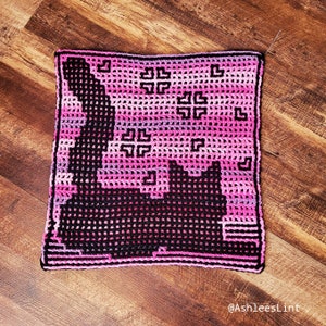 Könnte beinhalten: Ein quadratisches Kissen aus Häkelarbeit mit einem schwarzen Katzensilhouetten-Design auf einem rosa und violetten Ombre-Hintergrund. Das Kissen hat einen schwarzen Rand und ist aus einem strukturierten Garn gefertigt.
