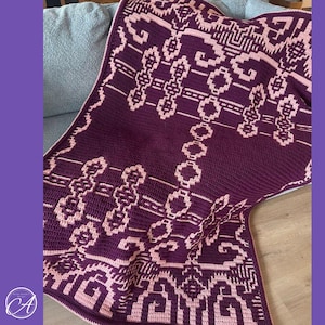 Könnte beinhalten: Eine gehäkelte Decke mit einem geometrischen Muster in Dunkelviolett und Hellrosa. Das Design besteht aus sich wiederholenden, komplexen Formen und Linien, die symmetrisch auf dem Stoff angeordnet sind und eine strukturierte, zweifarbige Oberfläche bilden.