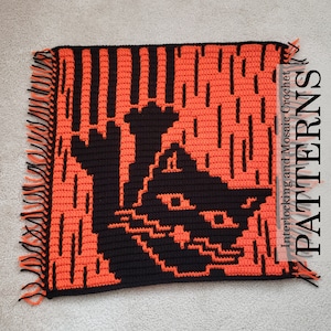 Op de afbeelding: Een zwart kattenhaakpatroon met een oranje achtergrond. Het patroon is gemaakt met behulp van in elkaar grijpende en mozaïekhaaktechnieken. De kat kijkt naar rechts en heeft een ondeugende uitdrukking. Het patroon is omlijst met zwarte franjes.