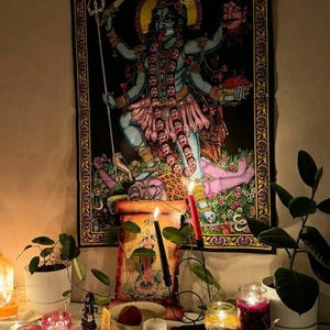 K&ouml;nnte beinhalten: Ein schwarzes Wandtuch, das die hinduistische G&ouml;ttin Kali mit mehreren Armen und einem wilden Ausdruck darstellt. Das Bild ist von einem dekorativen Rand umgeben. Die Szene umfasst Kerzen, Pflanzen und andere spirituelle Gegenst&auml;nde auf einer wei&szlig;en Oberfl&auml;che.